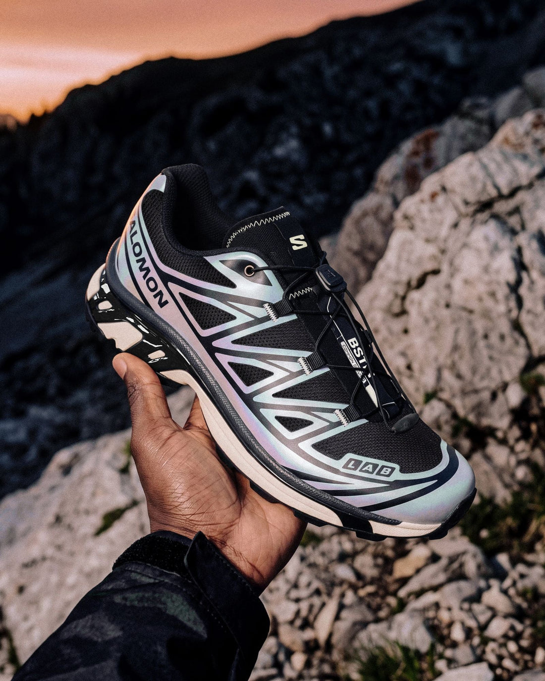 A hand holds up the BSTN x Salomon XT-6 5 AM with its iridescent silver upper against a sunrise over rocky mountains — official BSTN campaign image