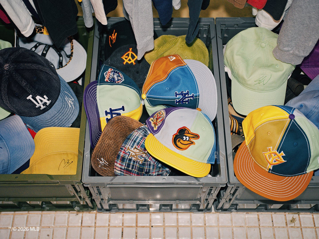 Crates full of New Era x Sean Wotherspoon caps under a rack of hanging clothes, including multicolor panel caps with Mets, Dodgers, Orioles and Giants logos plus corduroy and plaid styles — official New Era image
